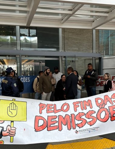 Imagem mostra protesto em frente a uma agência do Itaú, com uma faixa exposta em primeiro plano onde se lê "pelo fim das demissões"