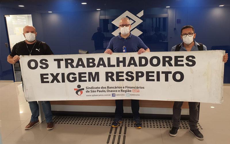 A partir da esquerda, os dirigentes sindicais Leonardo Diniz, Diego Pereira e Willame de Lavor protestam no Cenesp por resposta do banco à reivindicação por implementação do teletrabalho Três dirigentes sindiciais portam faixa com os dizeres "os trabalhadores exigem respeito". Ao fundo, o logo do Banco do Brasil