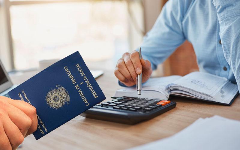 Imagem mostra, em primeio plano, uma mão de um homem branco segurando uma carteira de trabalho. Do outro lado da mesa, um homem de camisa azul clara e pele branca simula cálculos com uma calculadora e um caderno. os dois homens estão sentados em uma mesa de cor clara