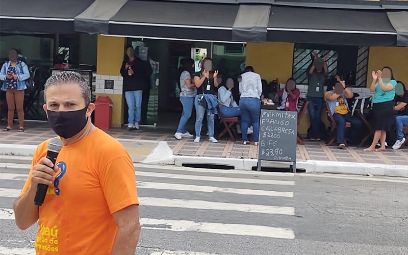 O dirgente sindical Sérgio Lopes, o Serginho, durante protesto que paralisou o CAT por conta de demissões A foto mostra o dirigente sindical Sérgio Lopes, o Serginho, com microfone na mão durante protesto contra demissões no Centro Administrativo Tatupé (CAT), doi Itaú. Ele é um homem branco, de cerca de 40 anos, pele clara, cabelos curtos e grisalhos; ao fundo bancários estão sentados em cadeiras na parte de fora de um restaurante, localizado do outro lado da rua; alguns deles estão apludindo o que Serginho diz