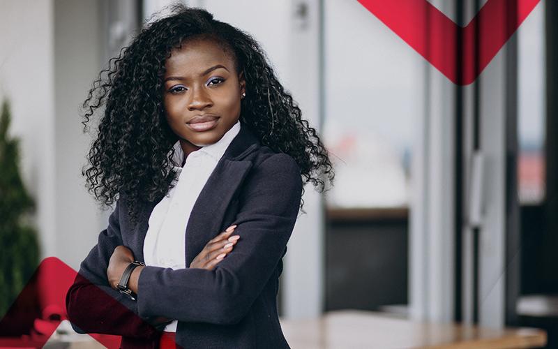 Imagem mostra uma mulher jovem negra, de cabelos compridos e soltos. Ela está com os braços cruzados e usa um terno cinza e uma blusa branca