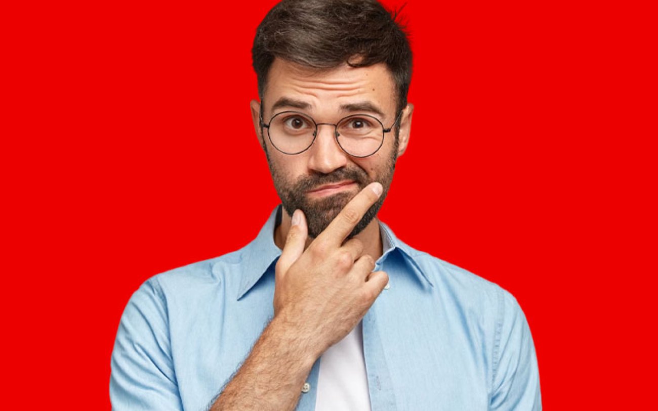 Imagem: Freepik Imagem com fundo vermelho mostra um homem jovem, branco, de barba e óculos,, vestindo camisa aberta e camiseta, com a mão no queixo e semblante de dúvida