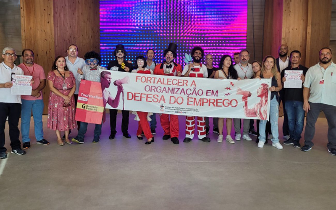 Imagem mostra dirigentes e funcionários do Sindicato dos Bancários de São Paulo durante mais uma etapa da campanha para denunciar a reestruturação em curso no Bradesco. Eles estão perfilados no saguão do Bradesco International Plaza e portam uma faixa onde se lê "Fortalecer a orgaização em defesa do emprego"