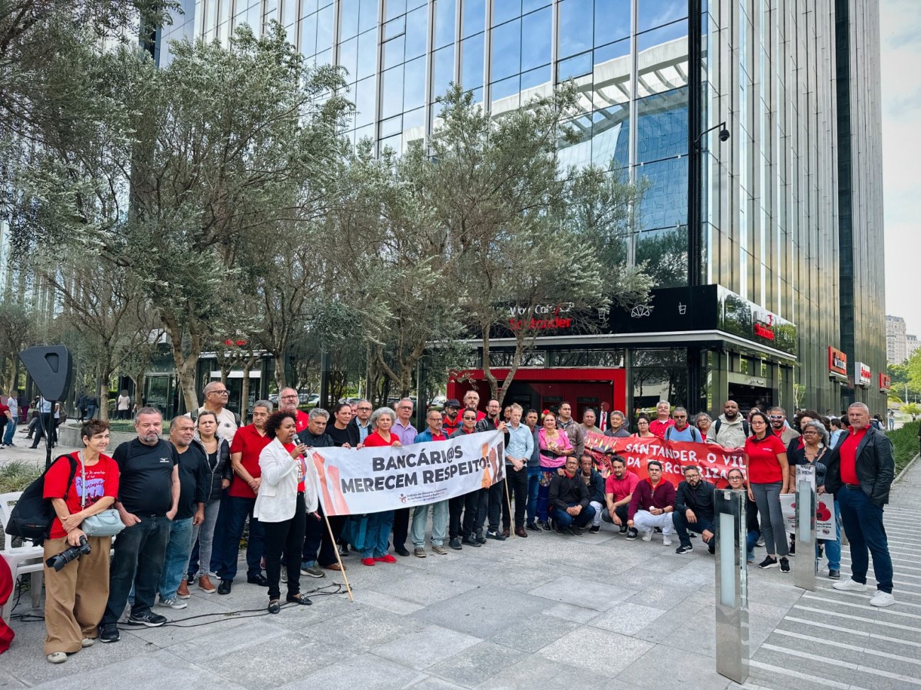 Protesto de trabalhadores contra as más práticas do Santander Fotografia mostra grupo de pessoas usando camiseta vermelha, portando faixa escrito bancários merecem respeito