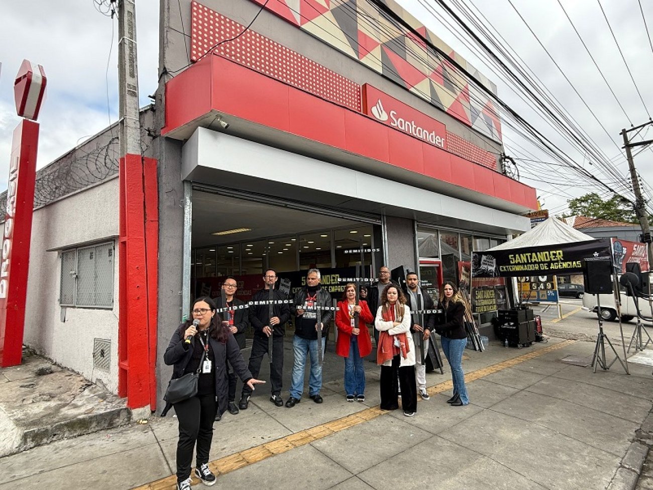 Imagem mostra dirigentes sindicais em frente a uma agência do santander, durante protesto contra o fechamento da unidade, os dirigentes estão de braços cruzados e lado a lado