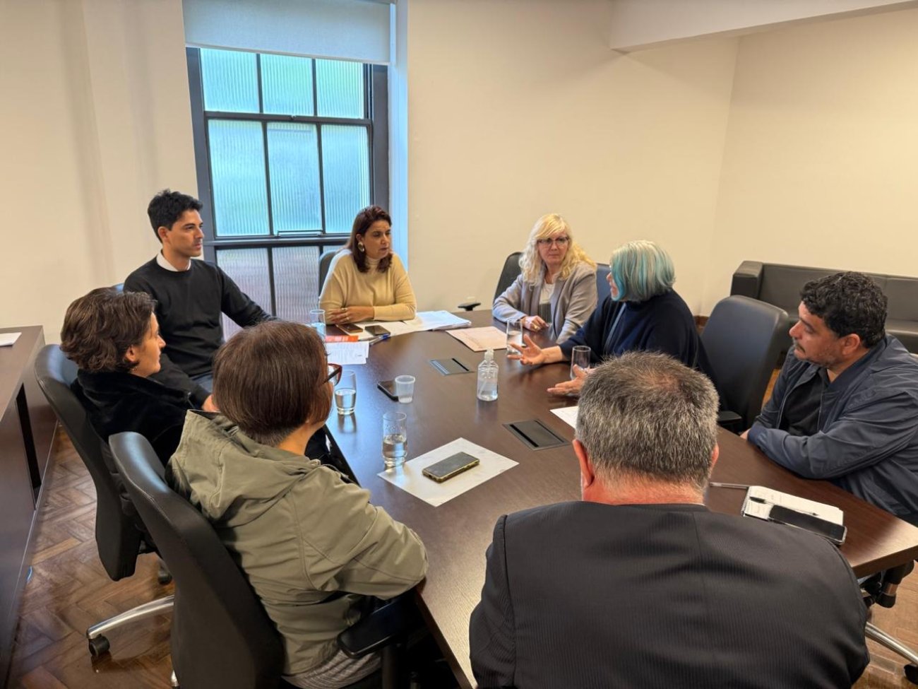 Imagem mostra reunião entre representantes do INSS e do Sindicato dos Bancários. São oito pessoas - cinco mulheres e três homens em volta de uma mesa oval marrom, em uma sala de parede branca