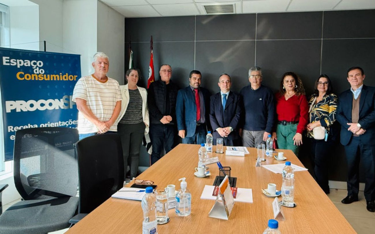 Imagem mostra participantes da reunião com o procon sobre o plano de saúde dos aposentados. Ao todos são nove pessoas. Três mulheres e seis homens. elas estão de pé e perfilados , em frente a ponta de uma mesa retangular. Ao lado deles há um banner onde se lê Procon espaço do consumidor