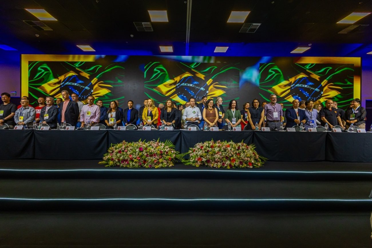 Imagem mostra palanque da abertura da 27ª conferência nacional dos bancários. vários dirigentes sindicais bancários estão perfilados lado a lado diante de uma mesa comprida. Um telão exibe três bandeira do Brasil, lado a lado