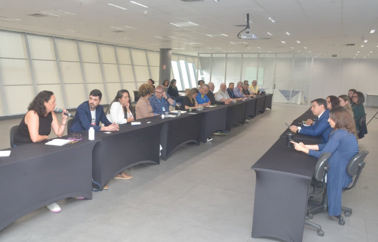 Imagem mostra sala onde ocorreu a reunião para discutir o Saúde Caixa. a imagem mostra 12 pessoas sentadas lado a lado em uma mesa comprida. Atrás dela há uma janela que ocupa toda a parede.