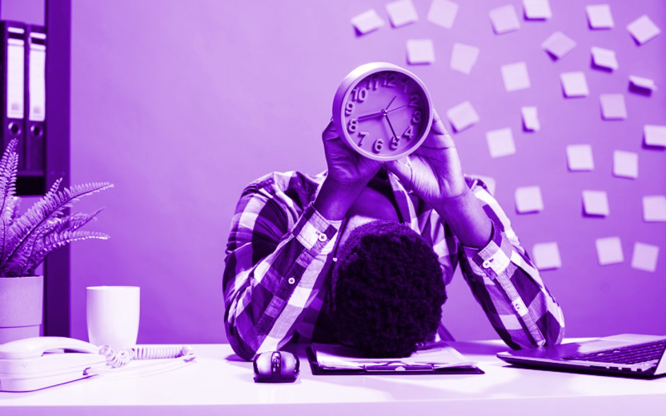 Imagem: Freepik Imagem de um trabalhador com cabeça apoiada na mesa e segurando um relógio
