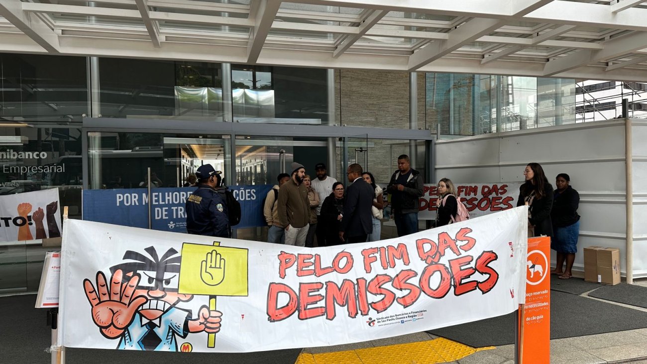 Imagem mostra protesto em frente a uma agência do Itaú, com uma faixa exposta em primeiro plano onde se lê "pelo fim das demissões"