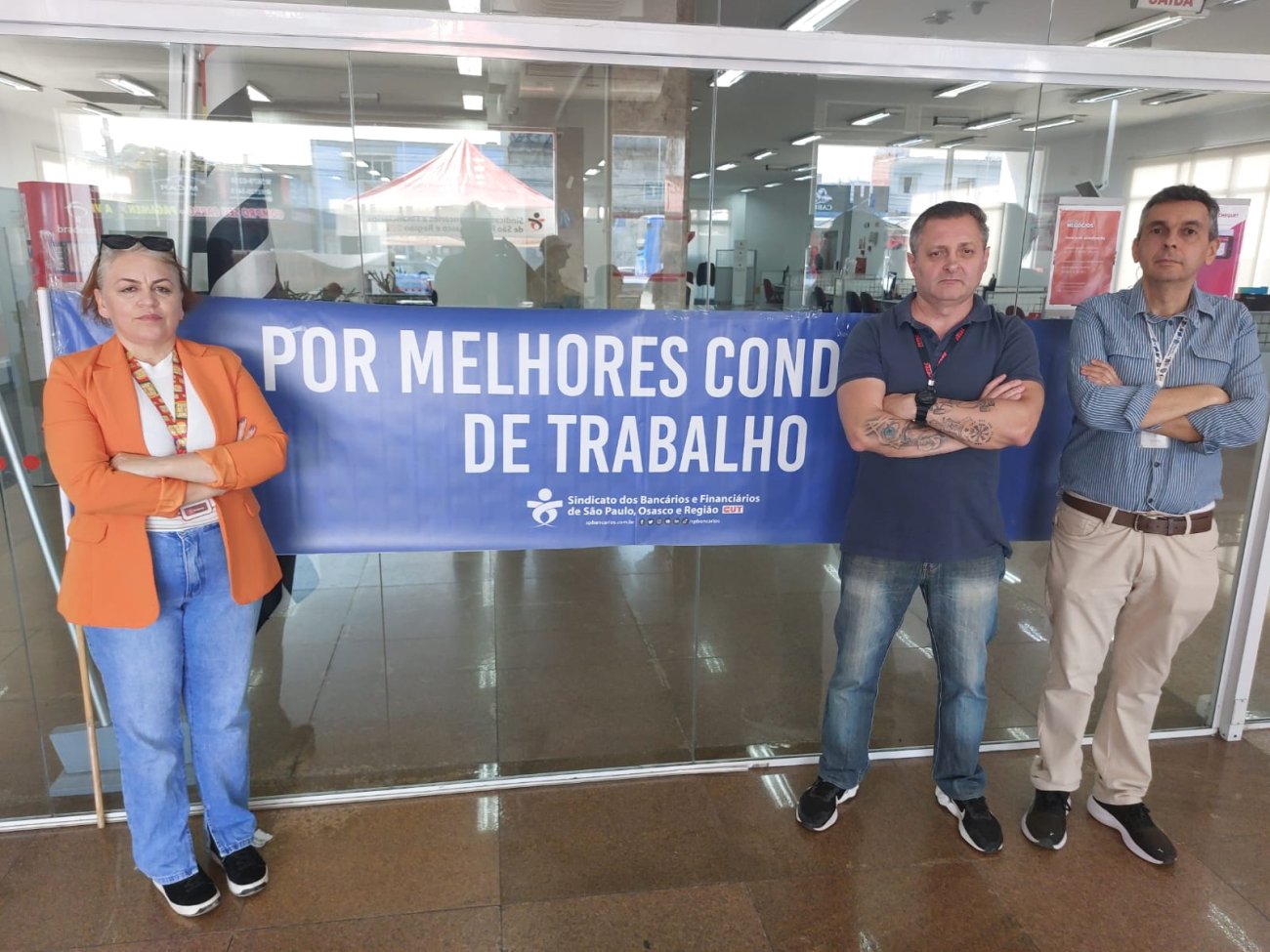 Imagem mostra dirigentes sindicais na porta de uma agência e ao lado de uma faixa onde se lê "por melhores condições de trabalho"