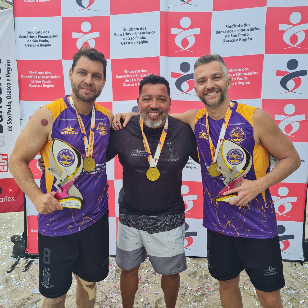Campeões da série Ouro do 1º Festival de Futevôlei dos Bancários Fotografia mostra 3 homens brancos de camiseta roxa e preta segurando troféu nas mãos