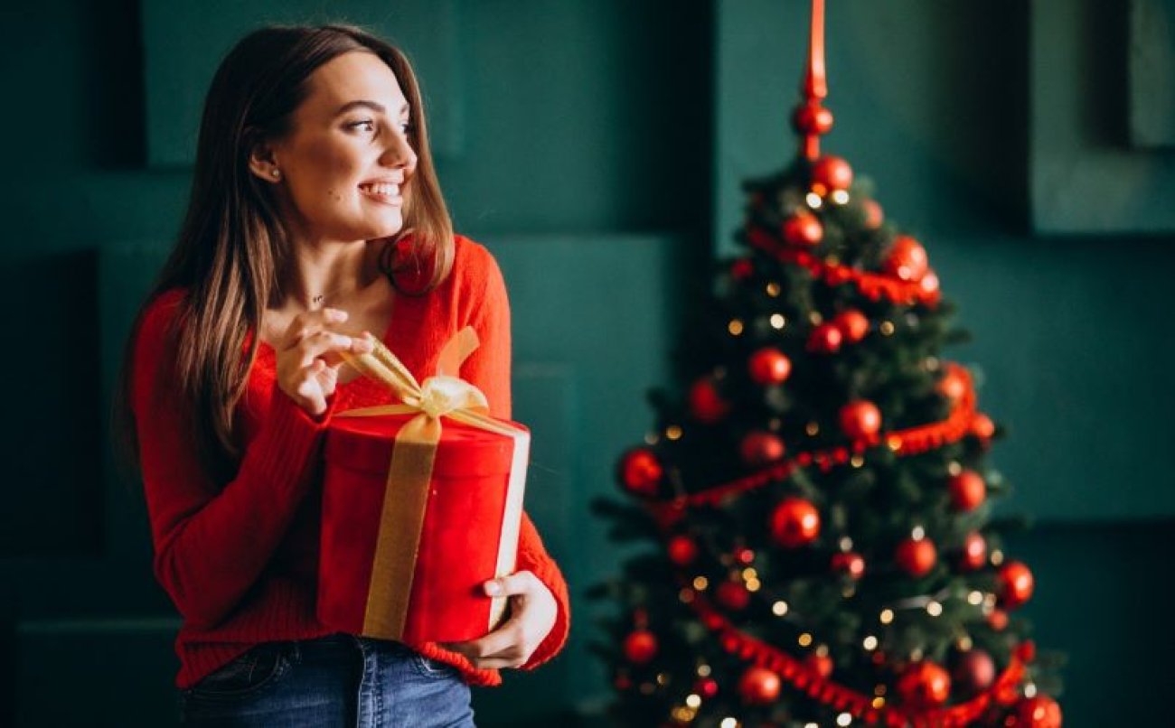 Imagem: Freepik Imagem de uma mulher segurando um presente, com árvore de natal ao fundo