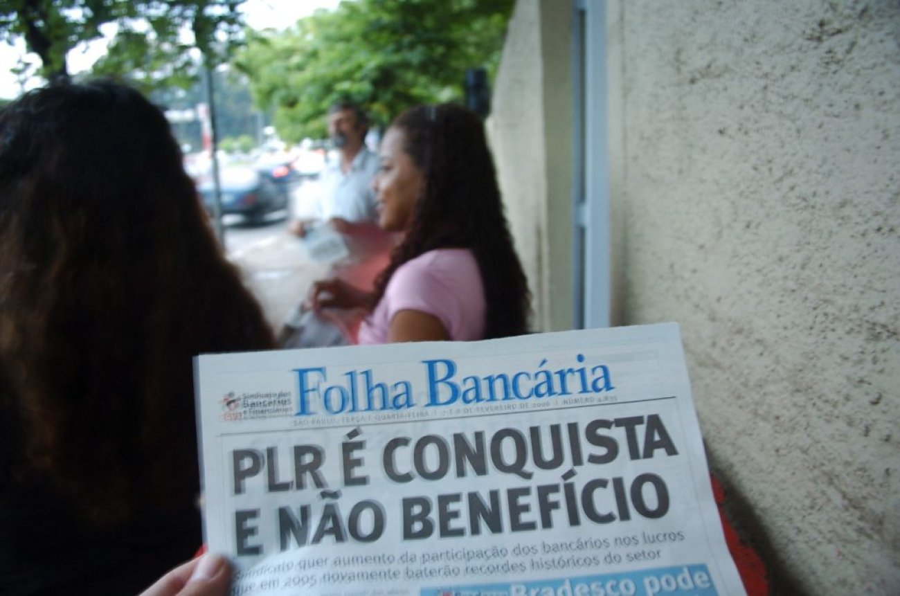 Não é benefício dado pelos bancos: há 30 anos bancários foram a primeira categoria a conquistar direito à PLR e garantida na CCT nacional (Crédito: Paulo Pepe/Seeb-SP) Fotografia mostra manchete de jornal falando sobre PLR é conquista, uma mulher negra de fundo de cabelos longos pretos e castanhos
