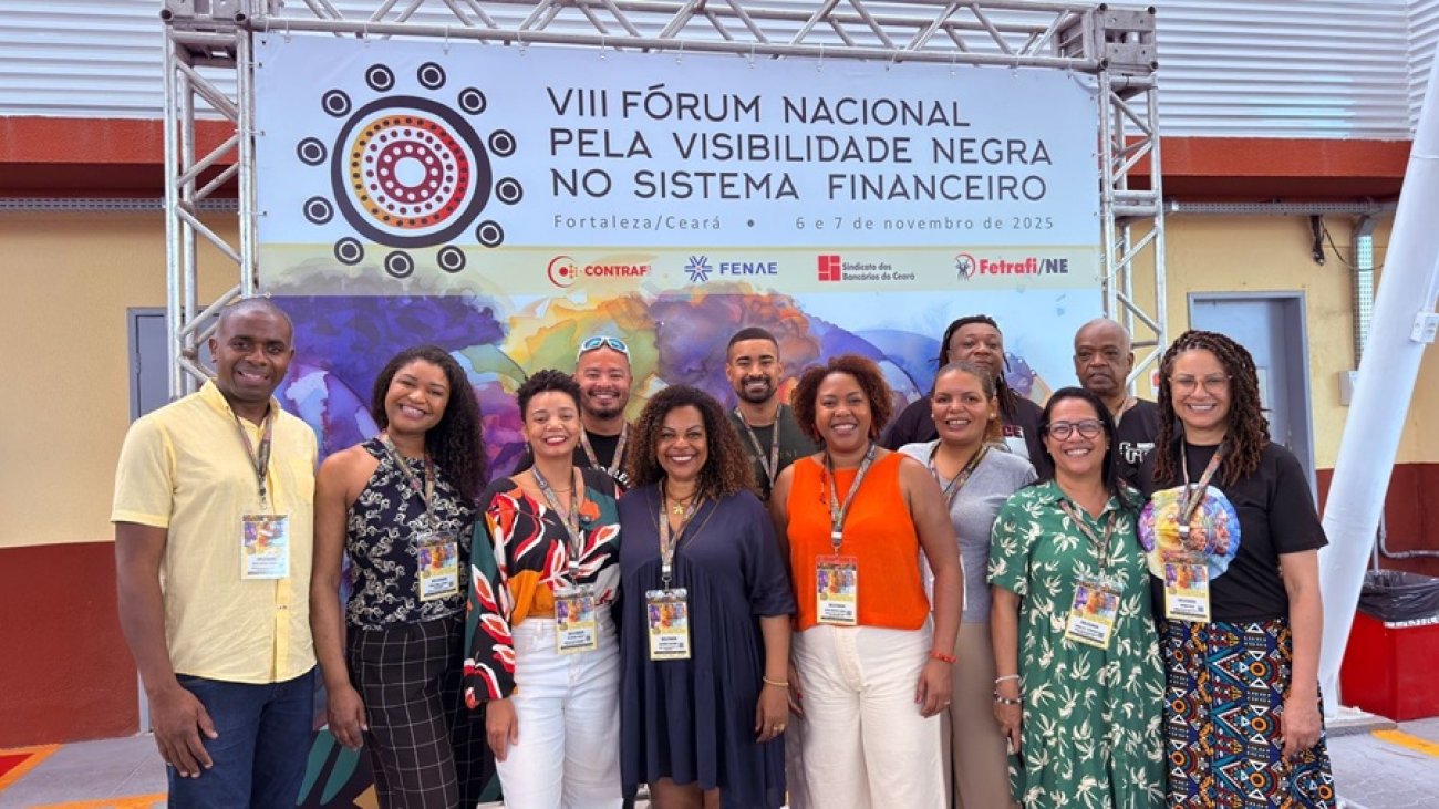 Imagem mostra membros do coletivo de combate ao racismo do Sindicato dos Bancáiros de São Paulo em frente a um painel do VIII Fórum Nacional pela Visibilidade Negra no Sistema Financeiro