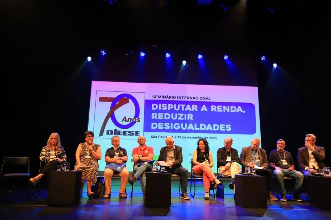 Imagem mostra representantes das centrais sindicais, sentados lado a lado em um palco, durante seminário do dieese. atrás deles há um painel onde se lê 70 anos Dieese. Seminário internacional. Disputar a renda, reduzir desigualdades