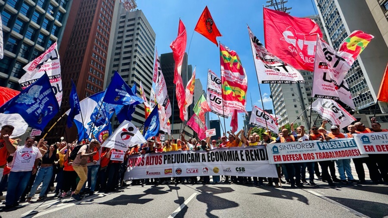 Imagem mostra manifestantes portando bandeiras de centrais sindicais e faixas durante protesto pela redução da taxa de juros