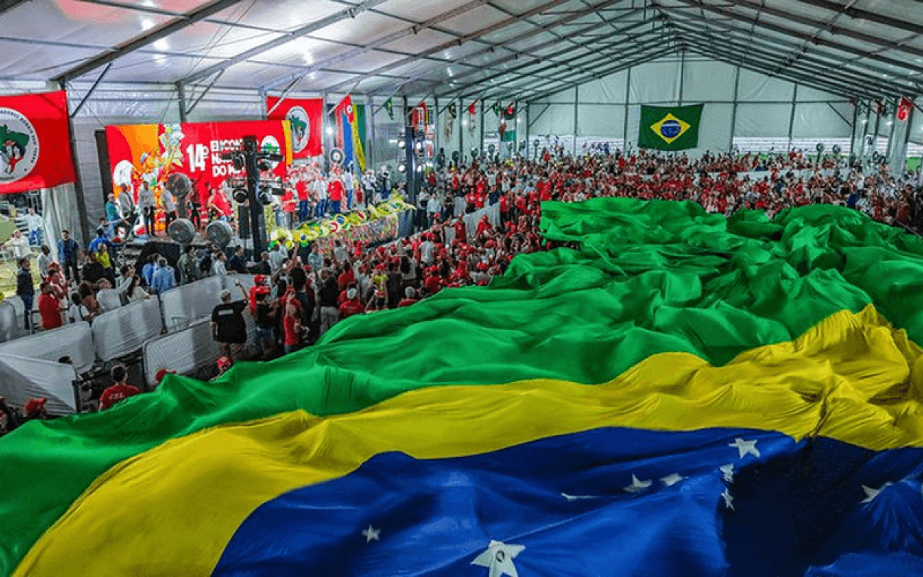 Foto mostra grande bandeira do Brasil sobre grande parte dos presentes em auditório onde ocorreu encerramento do encontro do MST