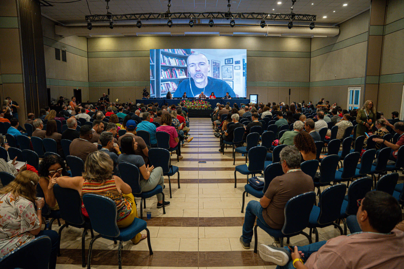 Foto do plenário do evento, tendo ao fundo a tela onde aparece o jornalista Jamil Chade