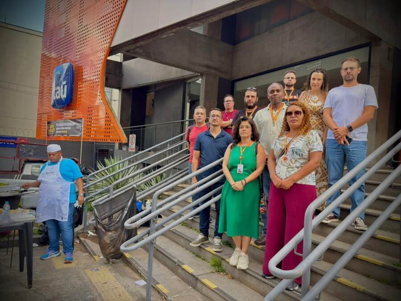 Imagem mostra dirigentes sindicais perfilados na escada em frente a uma agência do Itaú. Ao lado deles, um homem assa uma sardinha em uma grelha