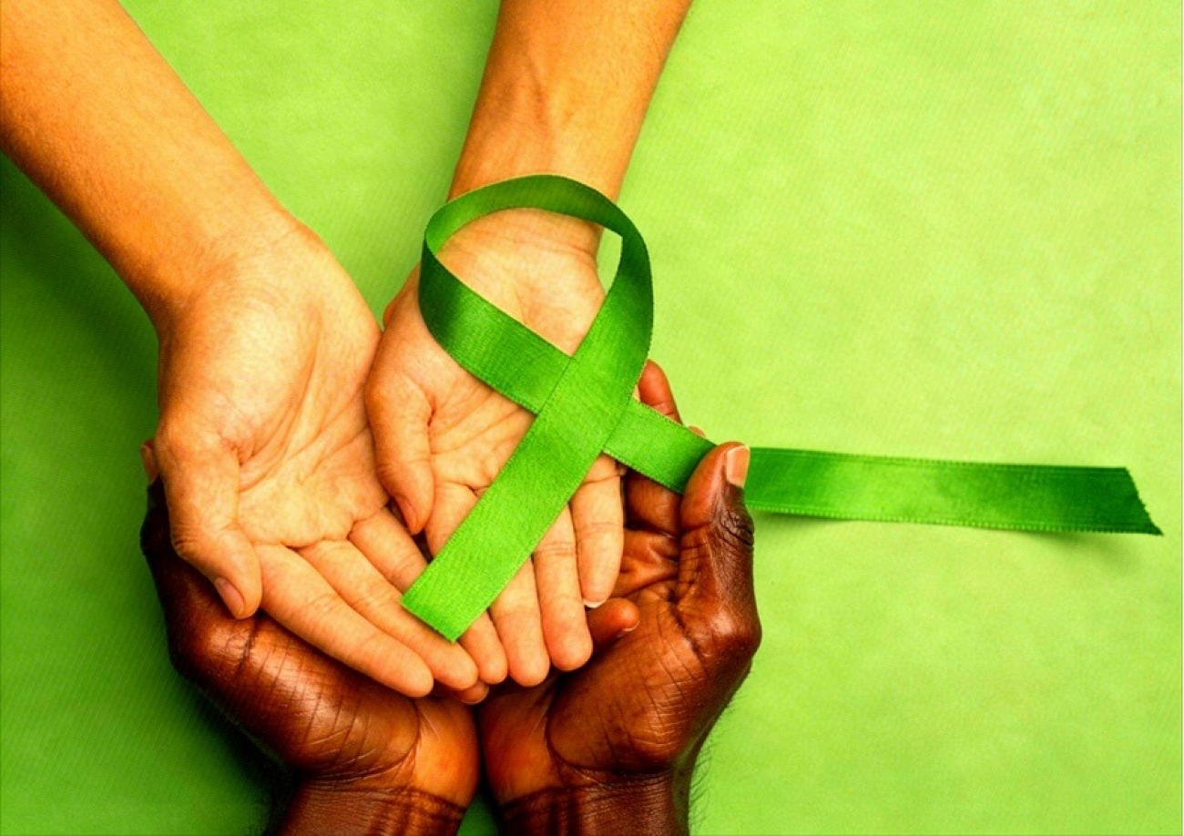 Imagem composta de um laço verde repousando sobre as palmas das mãos de uma pessoa branca. essas mãos estão sobre as palmas das mãos de uma pessoa negra. o fundo da imagem é verde