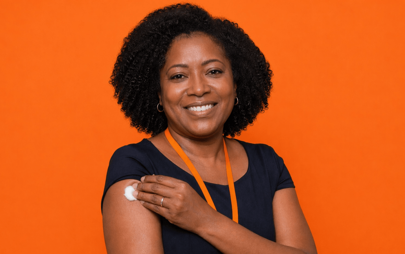 Fotografia de uma bancária negra, segurando um algodão no local da vacina, sorrindo. Ela veste preto e o fundo da imagem é laranja (Imagem: Manus.im)