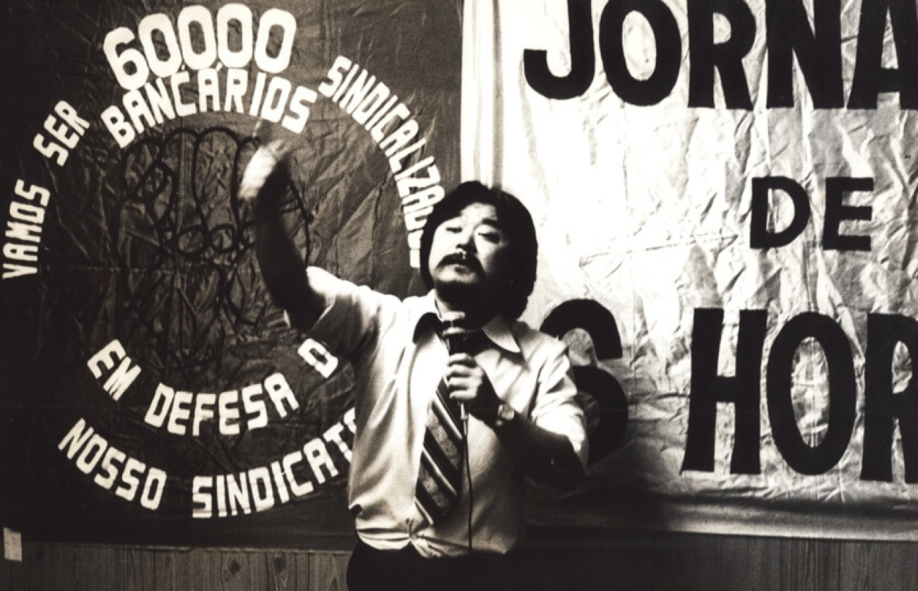 Imagem mostra Luiz Gushike, ex-presidente do Sindicato dos Bancários de São Paulo, Osasco e Região. ele é um homem oriental e, na foto, está na casa dos 30 anos. Veste camisa e gravata e fala ao microfone, com a mão esquerda levantada
