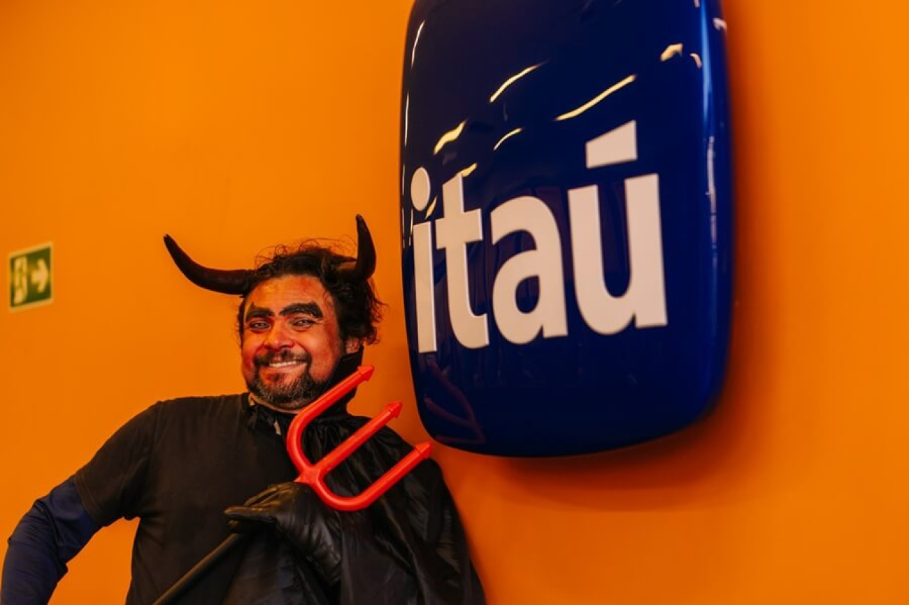 Imagem mostra homem fantasiado de diabo apontando um tridente para o logo do Itaú pendurado em uma parede pintada de laranja