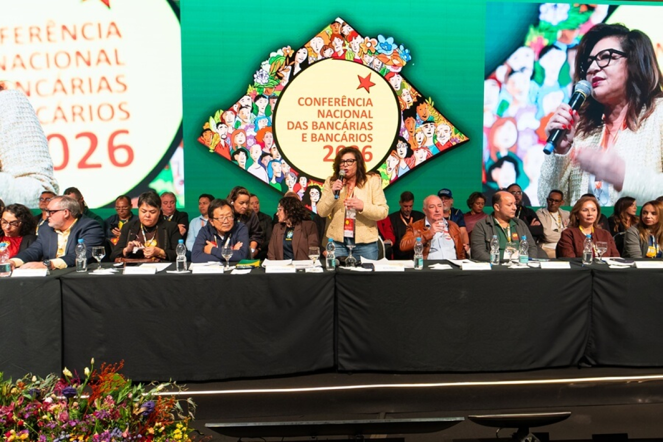 Imagem mostra Neiva Ribeiro de pé, no palco, falando ao microfone durante a abetura da 28ª Conferência Nacional dos Bancários