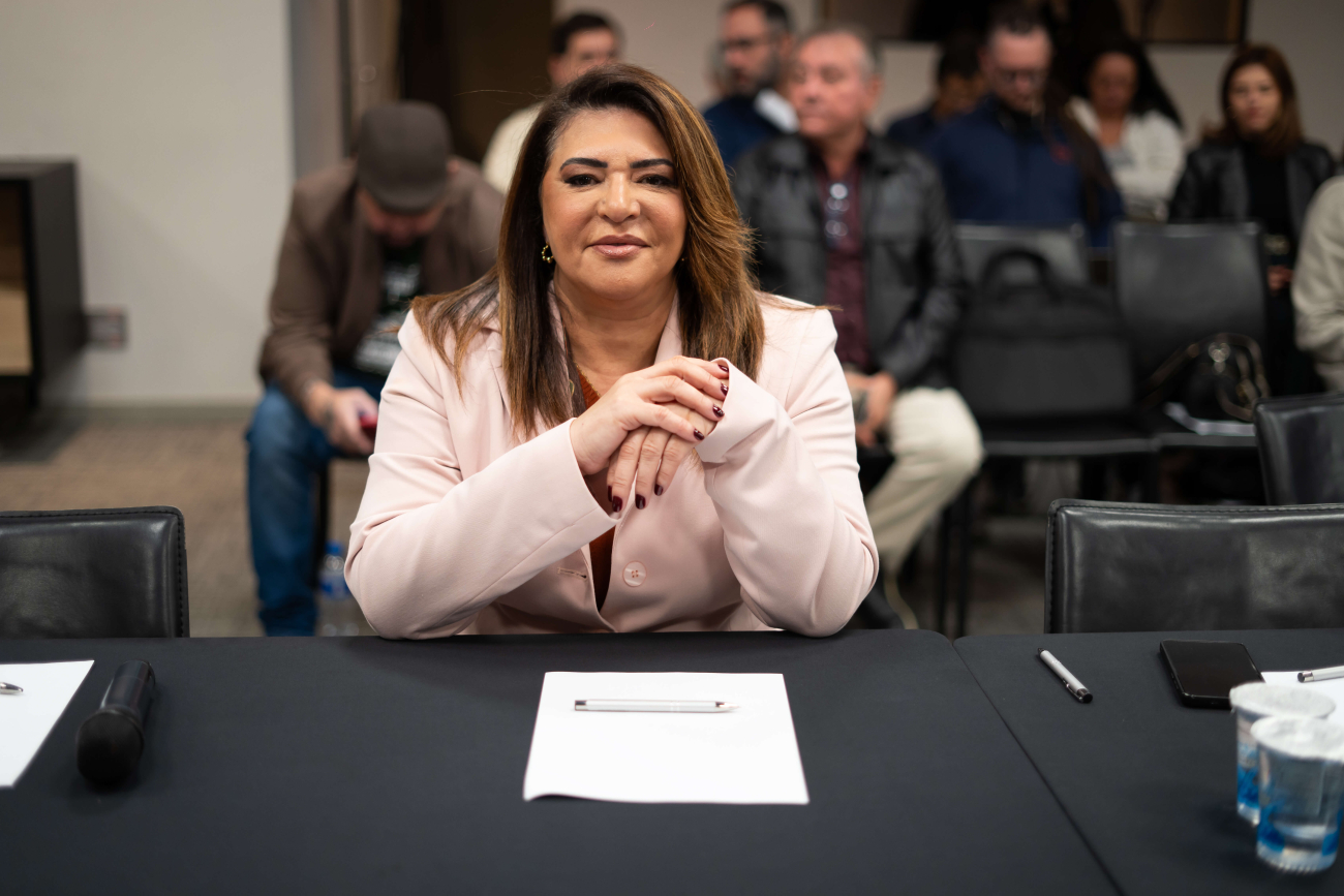 Neiva Ribeiro, presidenta do Sindicato dos Bancários, veste um blazer branco e usa óculos. Está sentada, com os braços apoiados em uma mesa, e dedos das mãos entrelaçados, na altura do peito