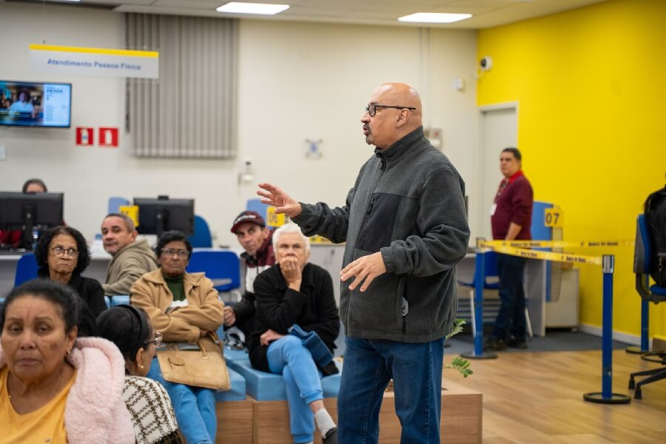 Imagem mostra o dirigente sindical João Maia (um homem branco, careca e de cerca de 50 anos), conversando com populares dentro de uma agência lotada do Banco do Brasil