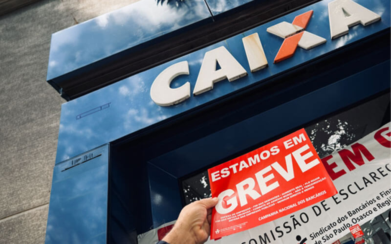 Imagem mostra uma mão segurando um adesivo onde se lê "greve", em frente à fachada de uma agência da Caixa