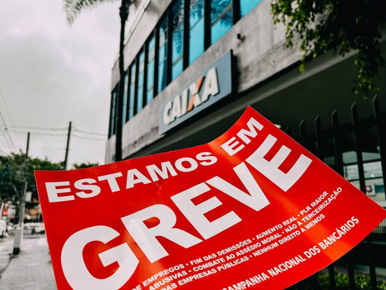 Imagem mostra um adesivo na cor vermelha onde se lê "Estamos em Greve" em letras brancas e, ao fundo, uma agência da Caixa Econômica Federal