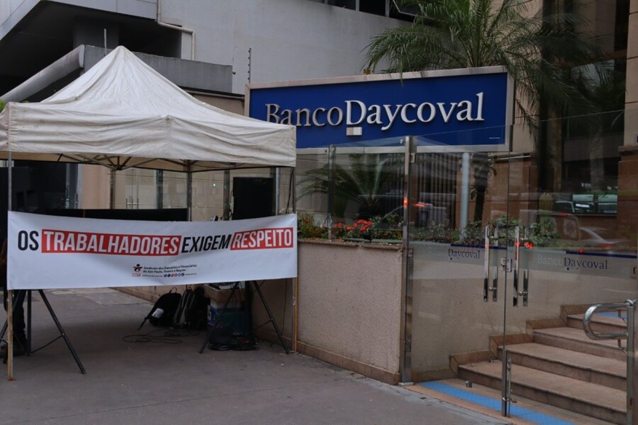 Imagem mostra faixa escrita "os trabalhadores exigem respeito" estendida em frente ao prédio do Daycoval e ao lado de uma placa com o logo do banco