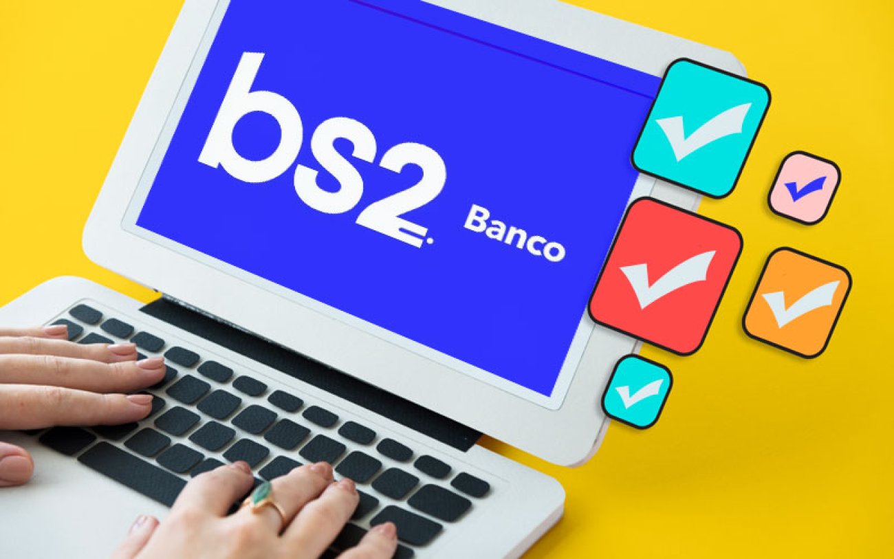 Arte com fundo amarelo mostra um notebook em cuja tela aparece o logo do banco BS2, ao lado de ícones de aprovação