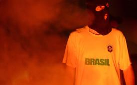 Imagem mostra um homem vestindo uma balaclava e uma camiseta amarela com o escudo da Confederação Brasileira de Furebol, onde se lê "Brasil", em meio a fumaça