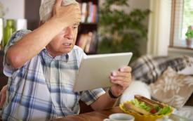 Imagem mostra um idoso sentado, com uma mão na testa, em sinal de preocupação, e a outra segurando um tablet