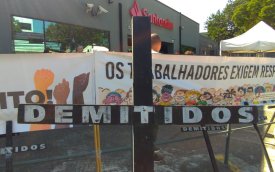 Imagem do protesto do Sindicato contra as demissões no Santander. A foto mostra uma cruz negra onde se lê "demitidos" e, atrás dela, faixas em frente à fachada do centro administrativo