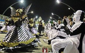 Fotografia mostra integrantes fantasiados de preto e branco ajoelhados ao lado de uma porta-bandeira da Vai-Vai