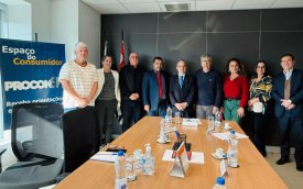 Imagem mostra participantes da reunião com o procon sobre o plano de saúde dos aposentados. Ao todos são nove pessoas. Três mulheres e seis homens. elas estão de pé e perfilados , em frente a ponta de uma mesa retangular. Ao lado deles há um banner  onde se lê Procon espaço do consumidor