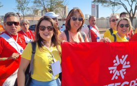 Fotografia mostra mulheres, uma de blusa vermelho e outra de amarela, segurando faixa vermelha