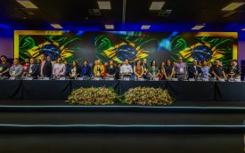 Imagem mostra palanque da abertura da 27ª conferência nacional dos bancários. vários dirigentes sindicais bancários estão perfilados lado a lado diante de uma mesa comprida. Um telão exibe três bandeira do Brasil, lado a lado