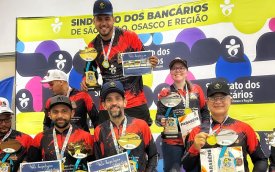 Fotografia mostra participantes do torneio de pesca com camisa colorida de vermelha, amarelo e preto segurando troféu nas mãos