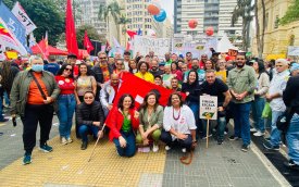 Homens e mulheres usando vermelho, alguns agachados e de fundo bandeiras de partidos e centrais sindicais