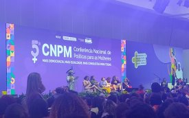 Imagem mostra plenário da 5ª conferencia nacional de política para as mulheres. a imagem foca o palco do evento enquanto discursa Sonia Guajajara, Ministra dos Povos Indígenas do Brasil