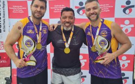 Fotografia mostra 3 homens brancos de camiseta roxa e preta segurando troféu nas mãos