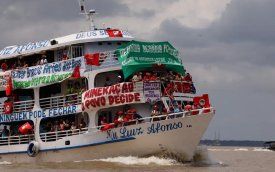 Imagem mostra um barco fluvial branco com detalhes em azul, de três andares e com bandeiras de movimentos sociais penduradas em seu casco