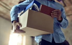 Imagem mostra homem vestido com uma camisa azul e segurando uma caixa com pertences, como se estivesse sendo demitido