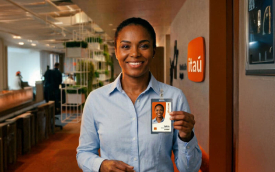 Imagem mostra uma mulher negra vestida com uma camisa azul, em um ambiente corporativa, segurando um crachá. ela sorri e, atrás dela, há fixado na parede um logo do itaú